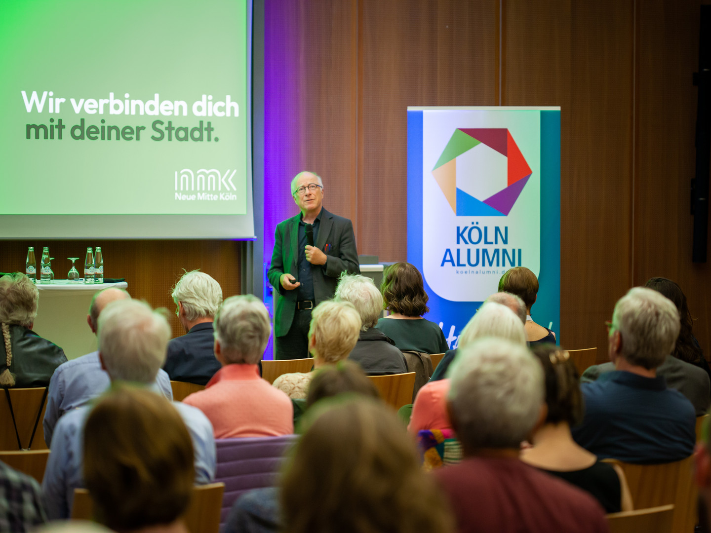 Paul Böhm zu Beginn seines Impulsvortrags vor dem Publikum des Abends. Im Hintergrund ist auf der linken Seite die Leinwand mit Böhms Präsentatiion zu sehen und rechts im Bild ein Aufsteller mit dem Logo von KölnAlumni.
