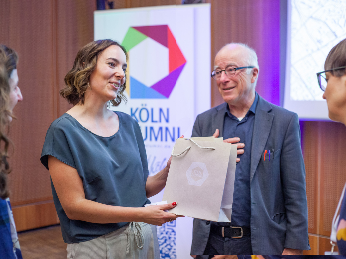 Anissa Vogel erhält ein Präsent von Carolin Bausum (Leiterin der Stabsstelle KölnAlumni). Im Hintergrund steht Paul Böhm mit einem Lächeln im Gesicht.
