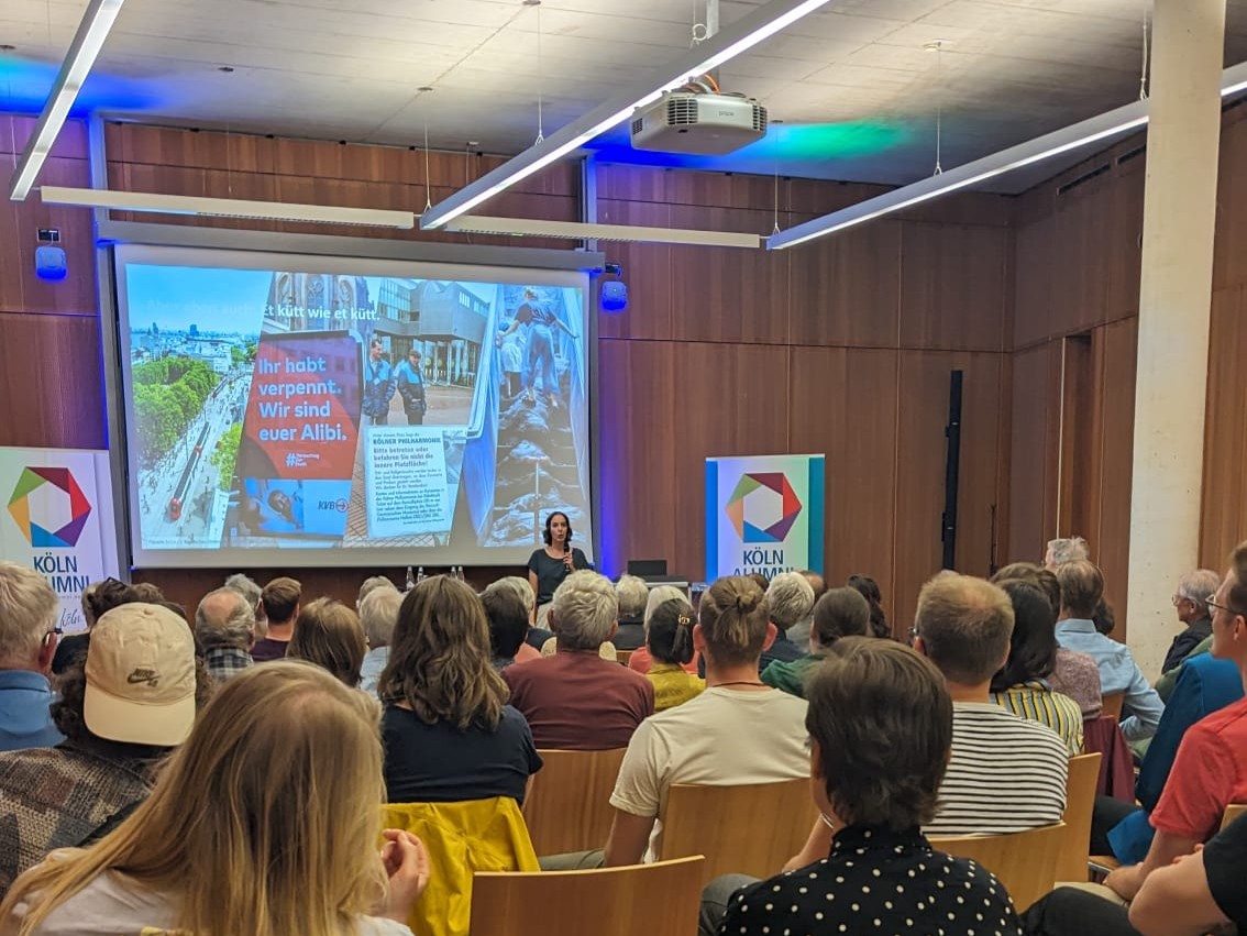 Anissa Vogel während ihres Vortrags, im Hintergrund ist ihre Powerpoint-Präsentation zu sehen. Das Publikum hört aufmerkam zu.